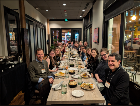 A group of UW Drama faculty, students, and alumni gathered around a long dinner table at a restaurant in Denver, smiling and celebrating their participation in the 2025 ASTR Conference. 