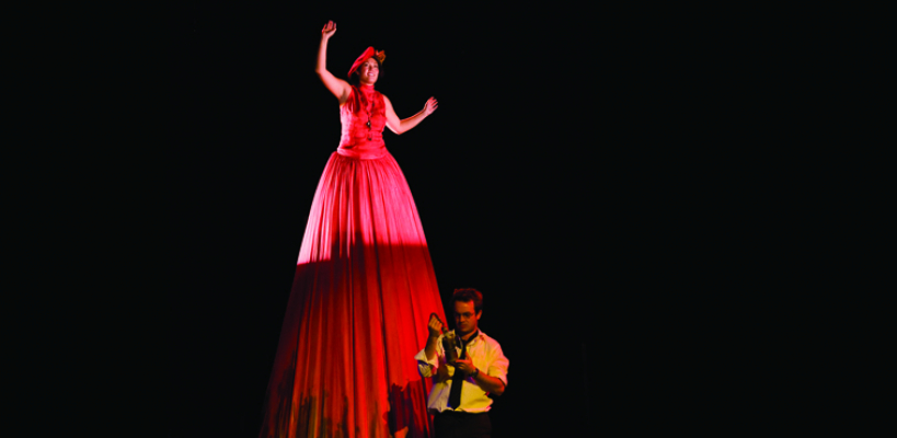 An actor appears in an extremely tall red dress in the play "Harp Song for a Radical"