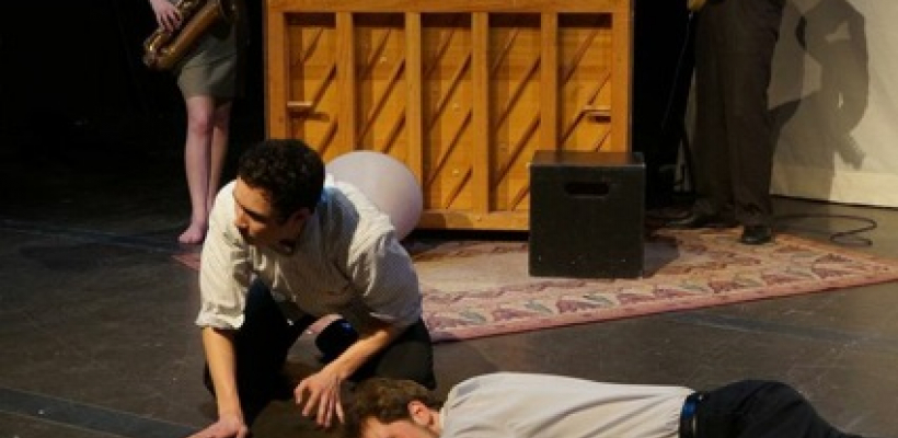 A man lays on top of a chalk outline, another crawls next to him. The musicians play in the background.