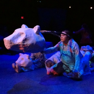 An actor on a blue-washed stage, operating a polar bear head puppet. Behind them, other actors represent sleeping cubs.