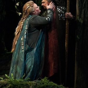 A Woman in blue leans in towards and rests her hand on a man's face who looks back at her amid a forestry surrounding.