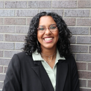 Woman wearing glasses and smiling stands in front of a grey brick wall.