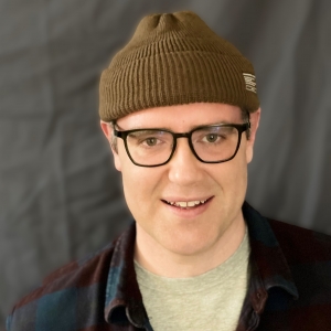 a person with a hat and glasses smiles in front of a dark background