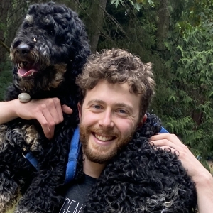 Ari Kaufman smiling and carrying a black curly-haired dog across his shoulders