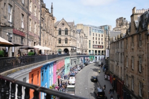 A curved road in Edinburgh lined with cars, people, and tan brick buildings