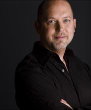 This is an image of Jeff Caldwell. He is staring and smiling at the camera. He is wearing a black shirt and folding his arms.