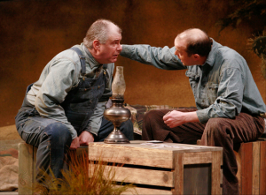 Two men in overalls sitting on bixes in hay talking to each other.