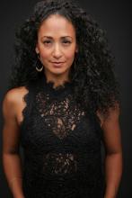 This is an image of Nikki Long. She is wearing a black shirt and standing with her hands to her side behind a black background. She is smiling and looking intensely at the camera. 