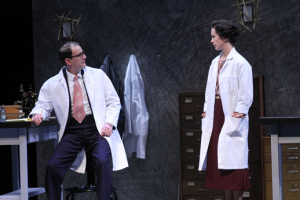 A man in a lab coat at a desk turns to talk to a woman in a labcoat.