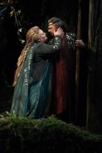 A Woman in blue leans in towards and rests her hand on a man's face who looks back at her amid a forestry surrounding.