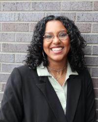 Woman wearing glasses and smiling stands in front of a grey brick wall.