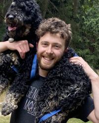 Ari Kaufman smiling and carrying a black curly-haired dog across his shoulders
