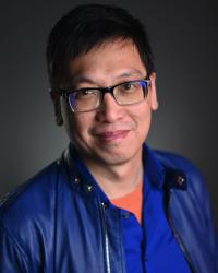 Czerton Lim in eyeglasses and blue leather jacket smiles at camera