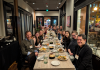 A group of UW Drama faculty, students, and alumni gathered around a long dinner table at a restaurant in Denver, smiling and celebrating their participation in the 2025 ASTR Conference. 