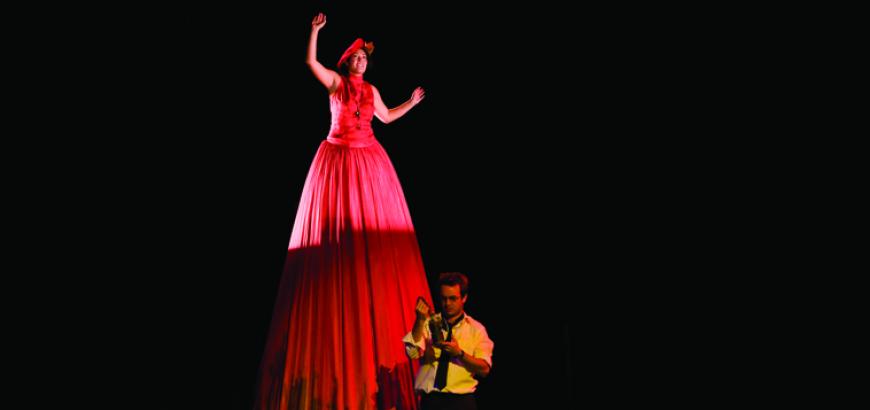 An actor appears in an extremely tall red dress in the play "Harp Song for a Radical"