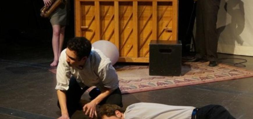 A man lays on top of a chalk outline, another crawls next to him. The musicians play in the background.