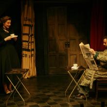 Mr. Peachum reads the newspaper while Mrs. Peachum smiles at him with a cup of coffee.
