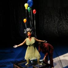 A strange faced woman has balloons come out from her while standing on a table.