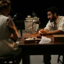 Two actors sit at a table, a woman facing away from the shot, a male leaning onto the table.