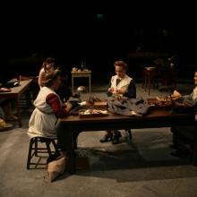Three women work at a table, while other women work behind them.