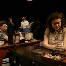 A woman sits at a table holding a glas, while other women sit behind her at other tables.