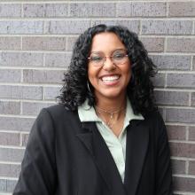 Woman wearing glasses and smiling stands in front of a grey brick wall.