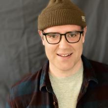 a person with a hat and glasses smiles in front of a dark background