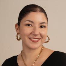 Headshot of Nour in front of a beige background. She wears a black shirt and gold jewlery.