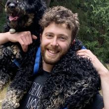 Ari Kaufman smiling and carrying a black curly-haired dog across his shoulders