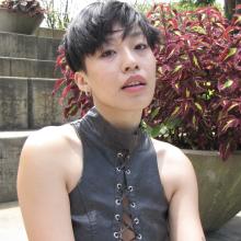 a light skinned Chinese-American nonbinary person with short black hair sitting in front of concrete steps and plants.