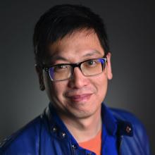 Czerton Lim in eyeglasses and blue leather jacket smiles at camera