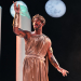 Actor in toga stands onstage with moon in background