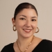 Headshot of Nour in front of a beige background. She wears a black shirt and gold jewlery.